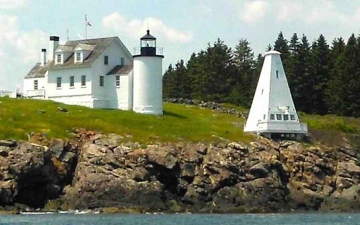 a white lighthouse building sits on green grass and rocks above water